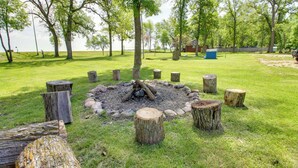 Un pozo de fuego rodeado de troncos de madera en un área cubierta de césped, con árboles y una vista distante de un lago.