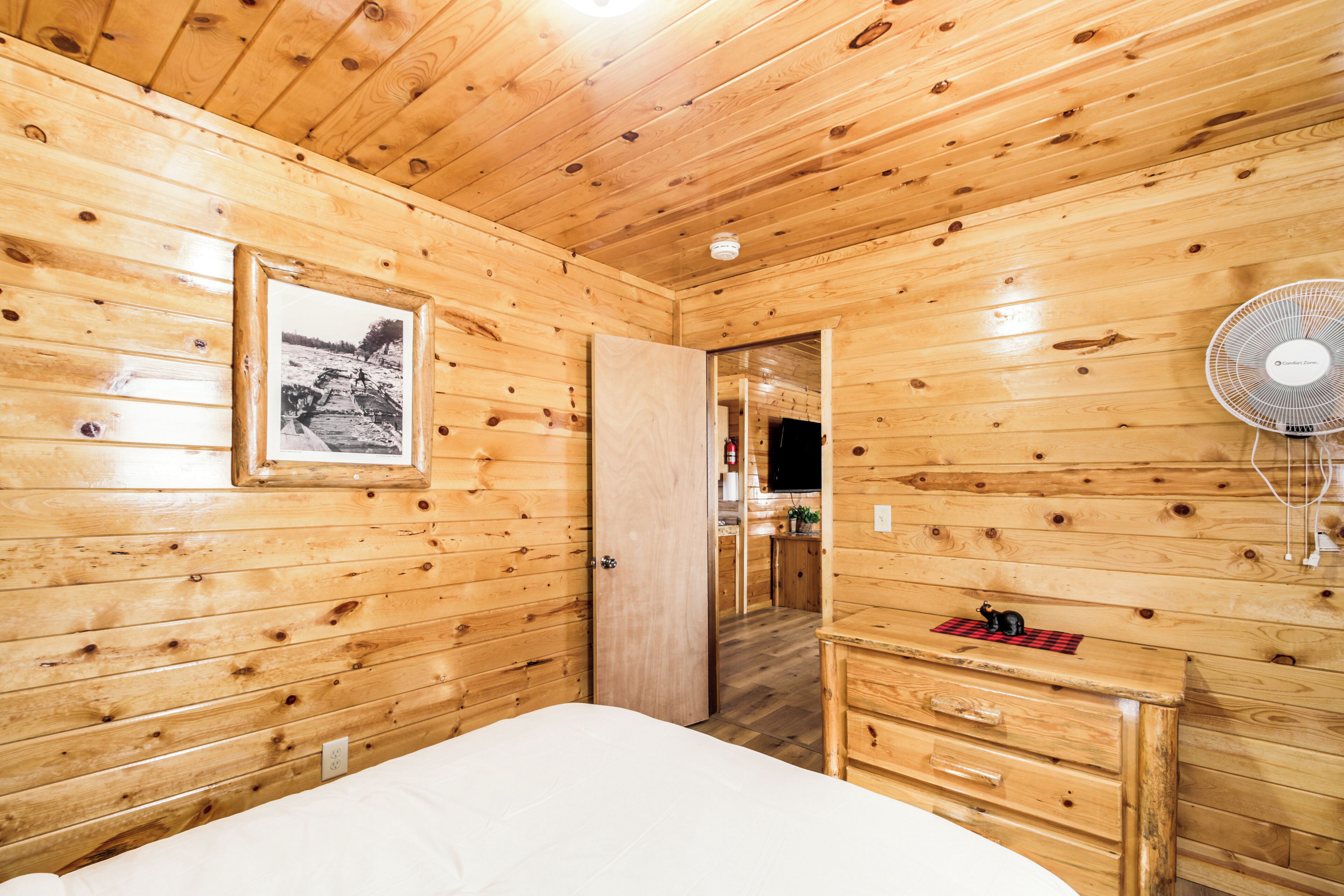 A wooden cabin room with a bed, a dresser, a framed picture on the wall, and a fan.