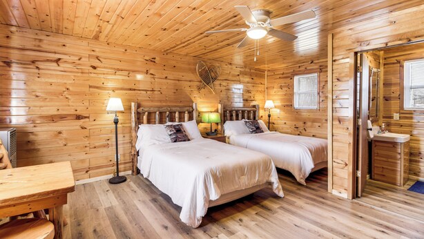 Una cabaña con dos camas, ventilador de techo, piso de madera y baño pequeño.