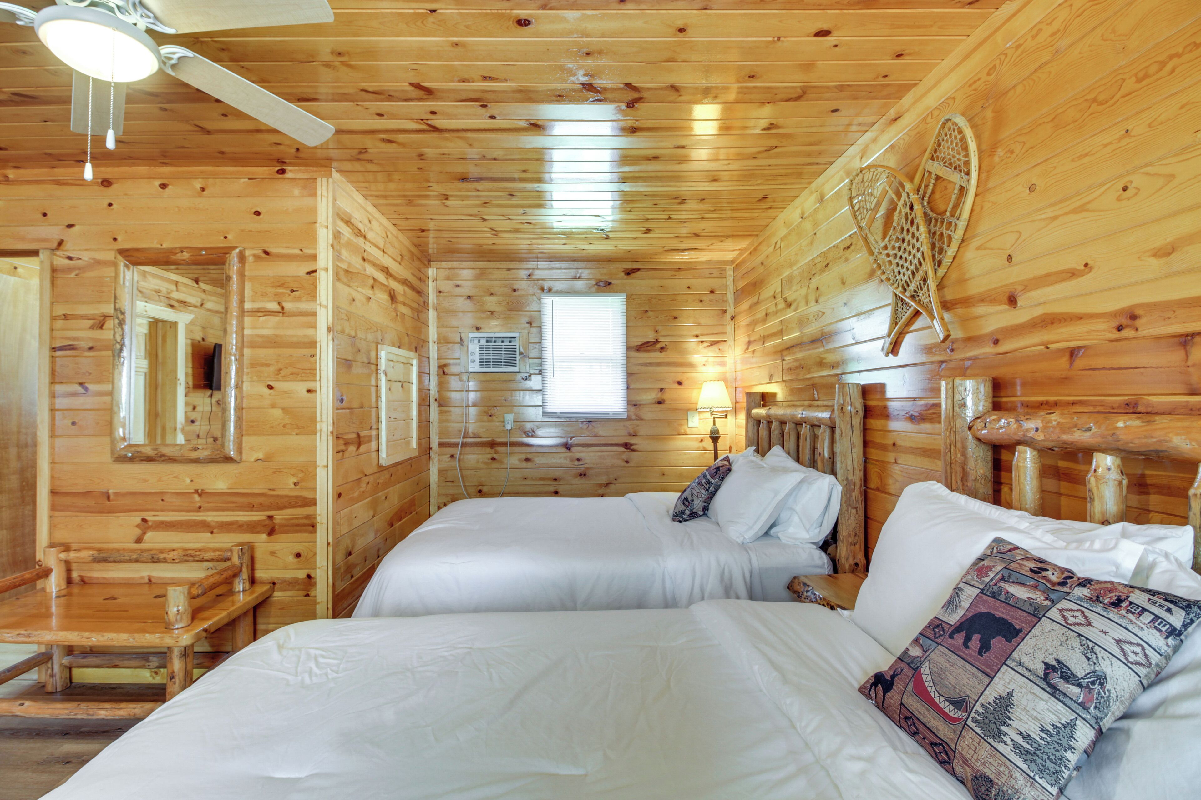 Una habitación tipo cabaña de madera con dos camas, un ventilador de techo, una ventana y un estante en la pared.