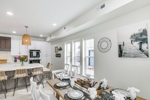 Une cuisine moderne avec des meubles blancs, un îlot central doté de tabourets de bar et un coin repas avec une table en bois prévue pour six personnes.