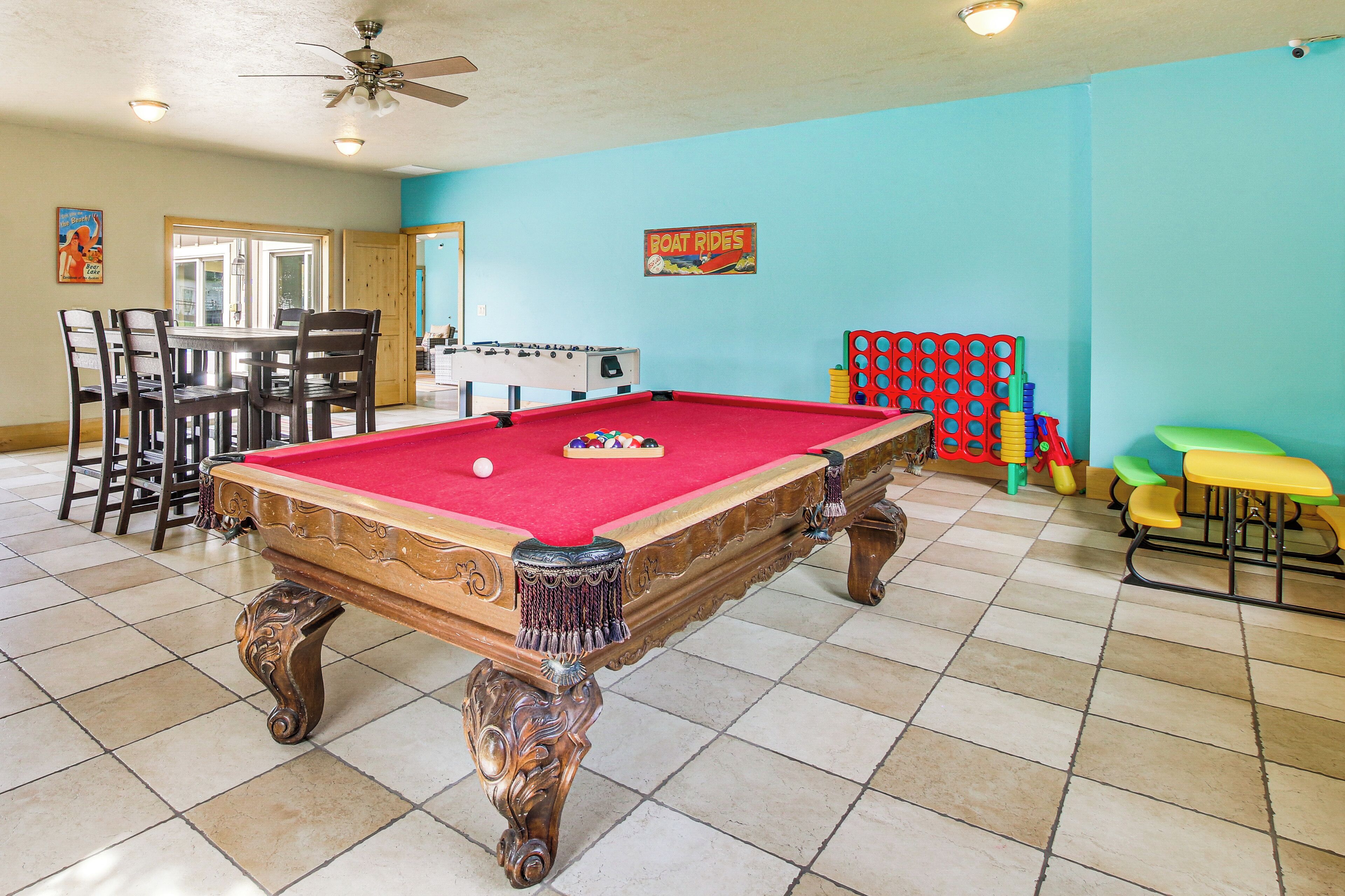 A pool table with a red felt surface, surrounded by wooden chairs and a foosball table.