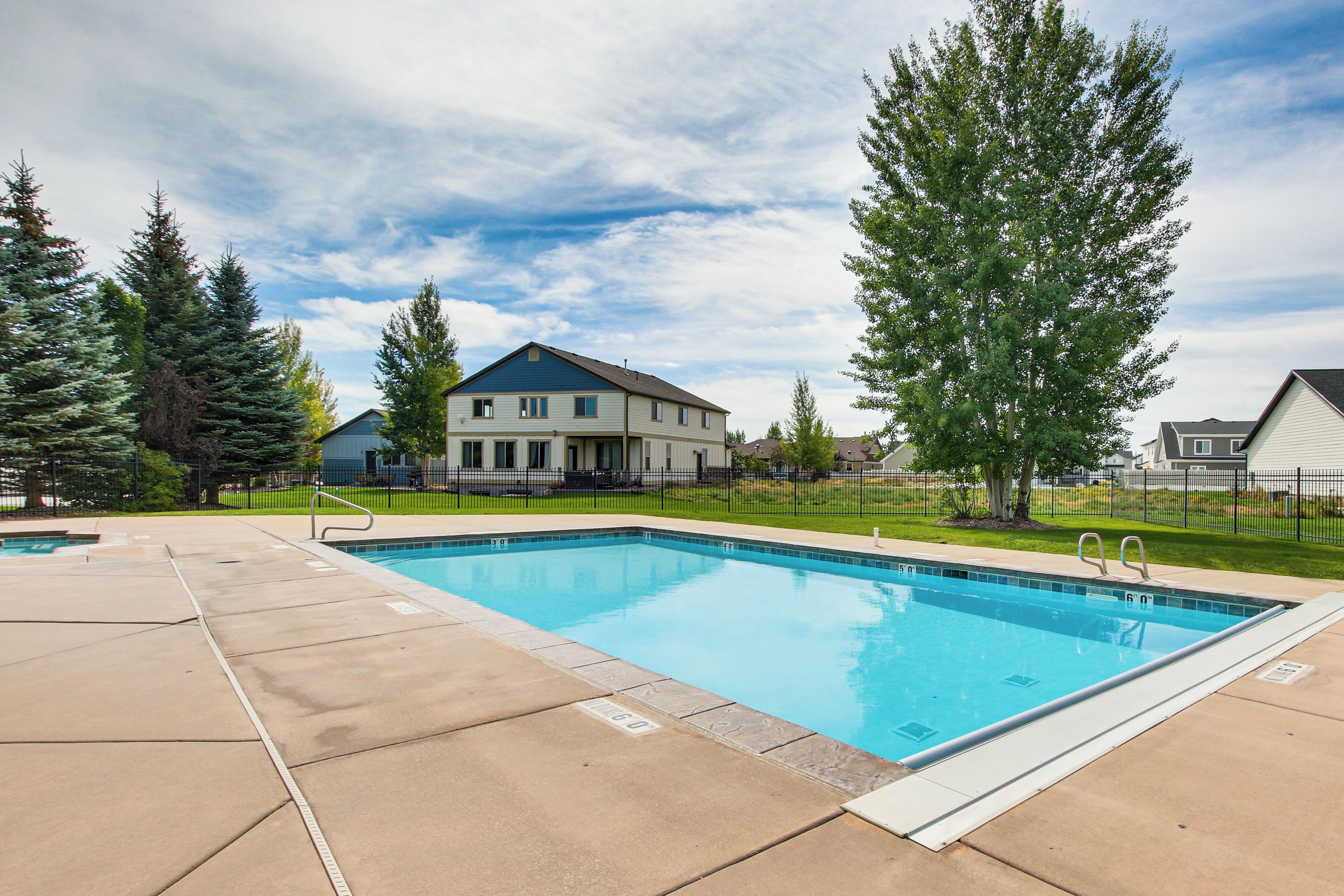 A rectangular swimming pool with a tiled deck, surrounded by a fence, and a large tree to the right.