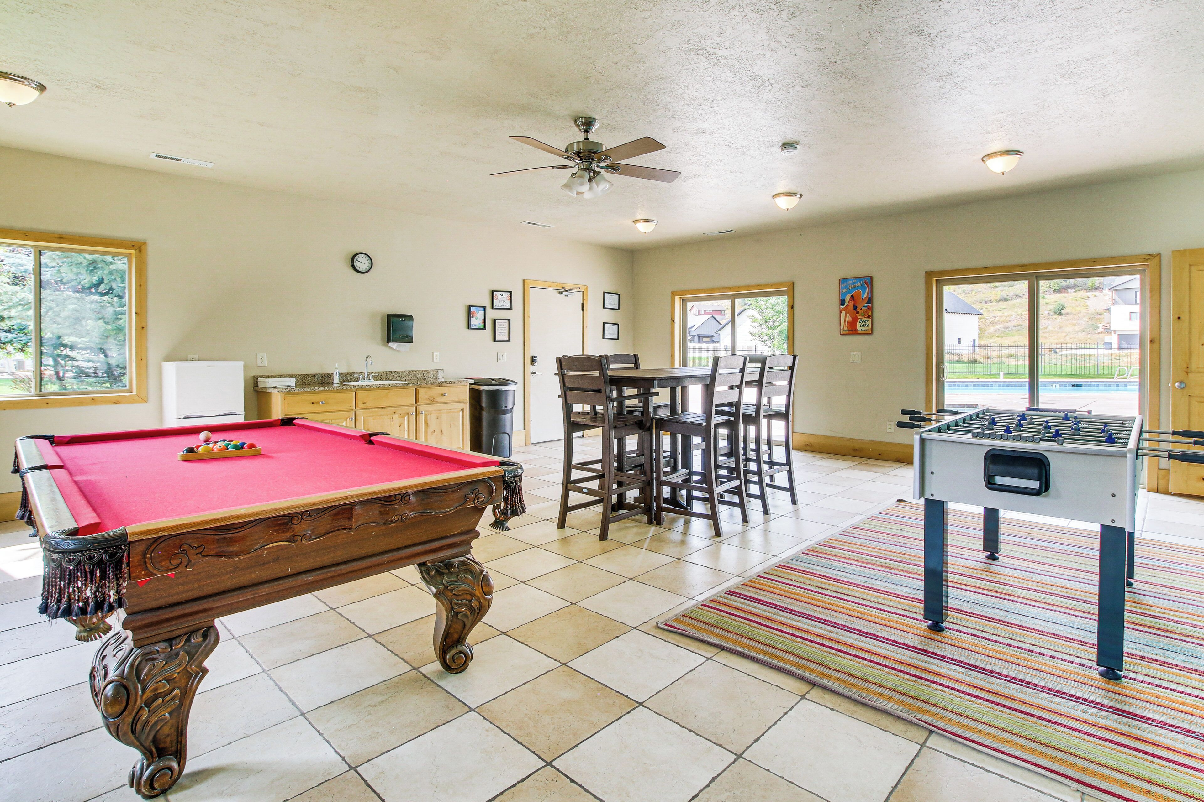 A pool table, dining table, and foosball table in a well-lit room with large windows.