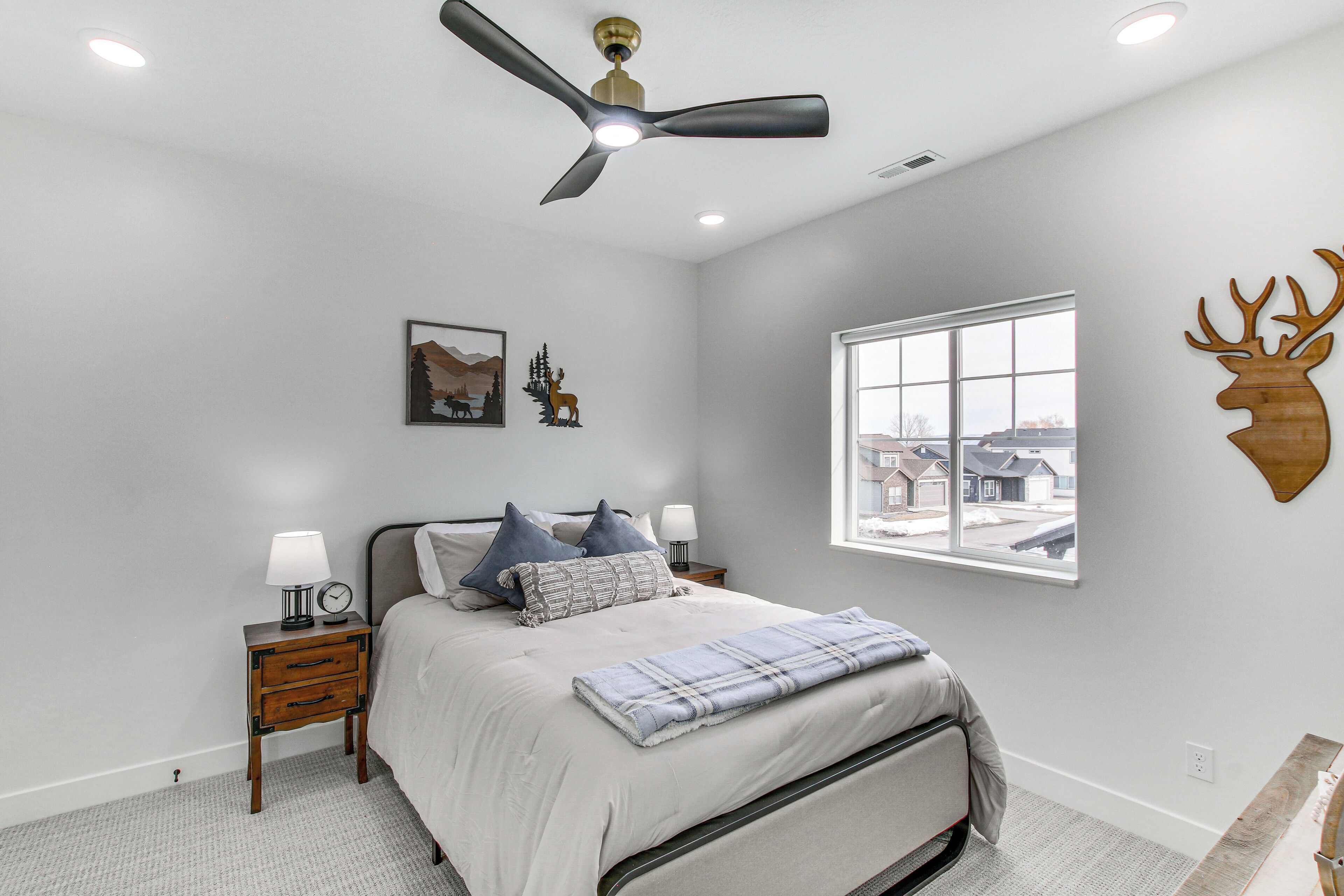 Une chambre à coucher comprenant un lit, des tables de chevet, un ventilateur de plafond, une fenêtre avec vue et des éléments de décoration murale, notamment une tête de cerf et un tableau représentant une montagne.