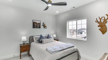 Une chambre à coucher comprenant un lit, des tables de chevet, un ventilateur de plafond, une fenêtre avec vue et des éléments de décoration murale, notamment une tête de cerf et un tableau représentant une montagne.