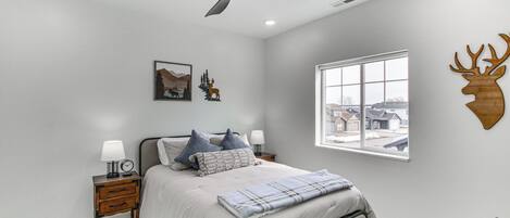 A bedroom with a bed, nightstands, a ceiling fan, a window with a view, and wall decorations including a deer head and a mountain painting.