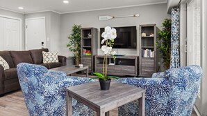 Una sala de estar moderna con un sofá marrón, sillones azules y blancos estampados, una mesa de centro de madera y un televisor sobre un mueble de madera.