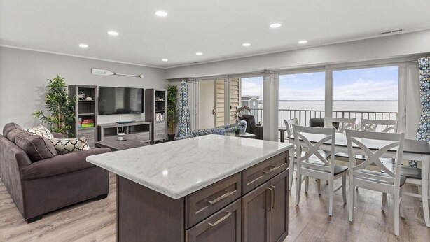 Un salón moderno con televisor grande, una zona de comedor con sillas blancas y vista al mar.