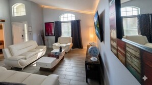 A living room with a sofa, armchairs, a coffee table, and a TV. There are windows with curtains, a door, and a wall-mounted globe.
