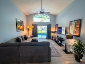 A living room with a sofa, a coffee table, a TV, a ceiling fan, and a large window with curtains. - Private Pool Close To Disney lg Hmslps 8 4 Bedroom Villa by RedAwning (Kissimmee)