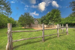 A rustic wooden cabin surrounded by a wooden fence in a grassy area with trees. - Town Creek Cabin Walk to Main St. (Fredericksburg)