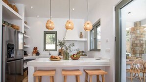 A modern kitchen with a central island, three wooden stools, hanging wicker pendant lights, and a stone wall visible through a glass door. - Villa Elaia is the perfect retreat for those seeking a luxurious a getaway. (Rhodes)