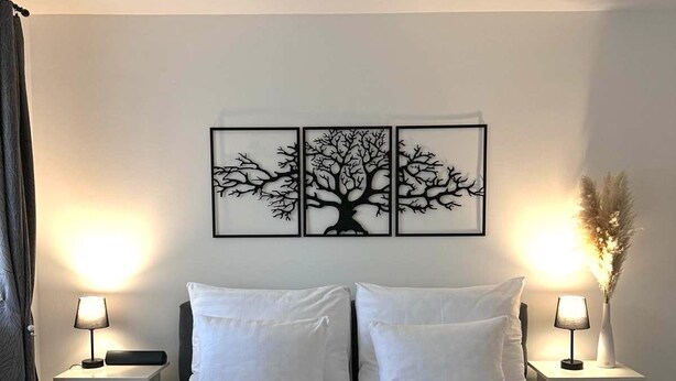 Une chambre d’hôtel avec un grand lit, deux tables de chevet avec des lampes, une œuvre d’art représentant un arbre accrochée au mur, et une fenêtre avec des rideaux.