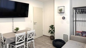 Un salon moderne doté d’un téléviseur à écran plat, d’un coin repas avec une table et des chaises, d’un panier noir pour animaux de compagnie et d’une petite étagère ornée d’objets décoratifs.