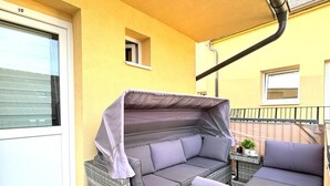 Un espace extérieur aménagé, abrité, avec un canapé et des fauteuils, une petite table et une pancarte indiquant « MAISON » posée sur le sol.