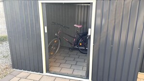 Un abri de jardin en métal avec une porte, un vélo à l’intérieur et un sol pavé de briques.
