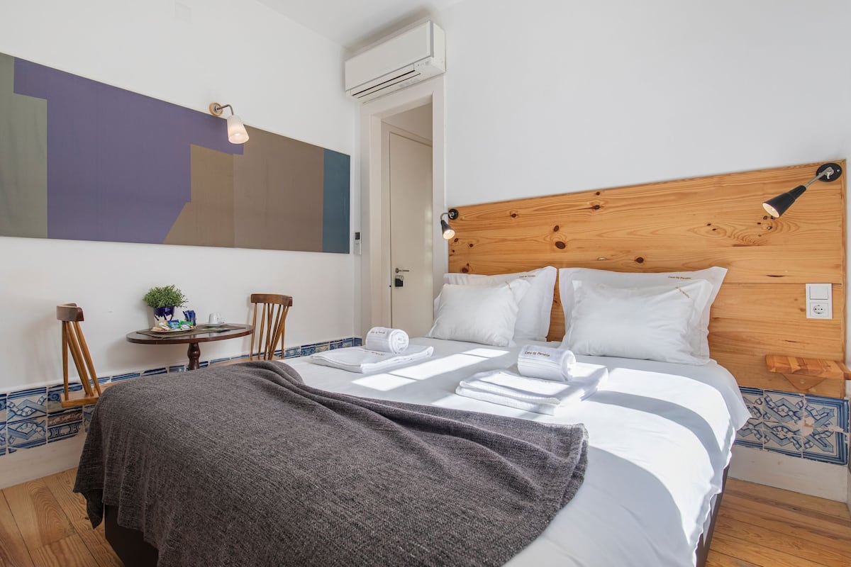 Un lit bien fait, avec du linge de lit blanc, une couverture grise et une tête de lit en bois. On y trouve une petite table sur laquelle sont posés une plante et une lampe.