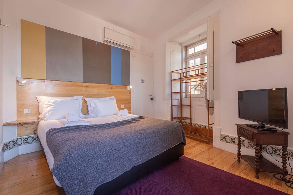 Une chambre avec un grand lit, une tête de lit en bois, un téléviseur posé sur un meuble en bois et une fenêtre donnant sur l’extérieur.