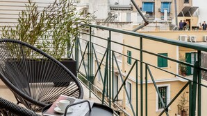 A balcony with two black wicker chairs, a small round table, and a potted plant. The balcony overlooks a residential area with buildings and balconies.