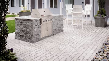 An outdoor patio with a stone grill station, white chairs, and a building with multiple doors in the background.