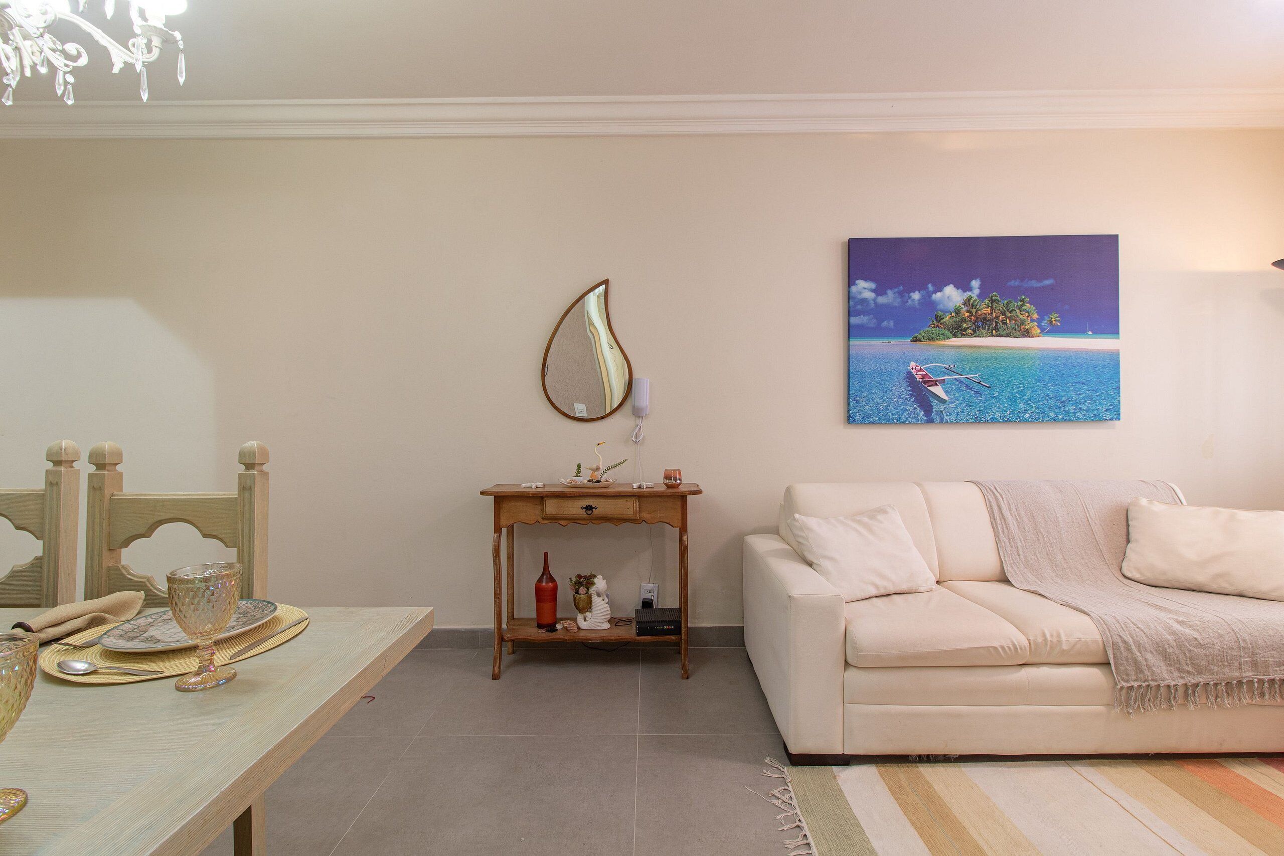 Un salon avec un canapé beige, une table basse en bois et un tableau représentant une île tropicale.