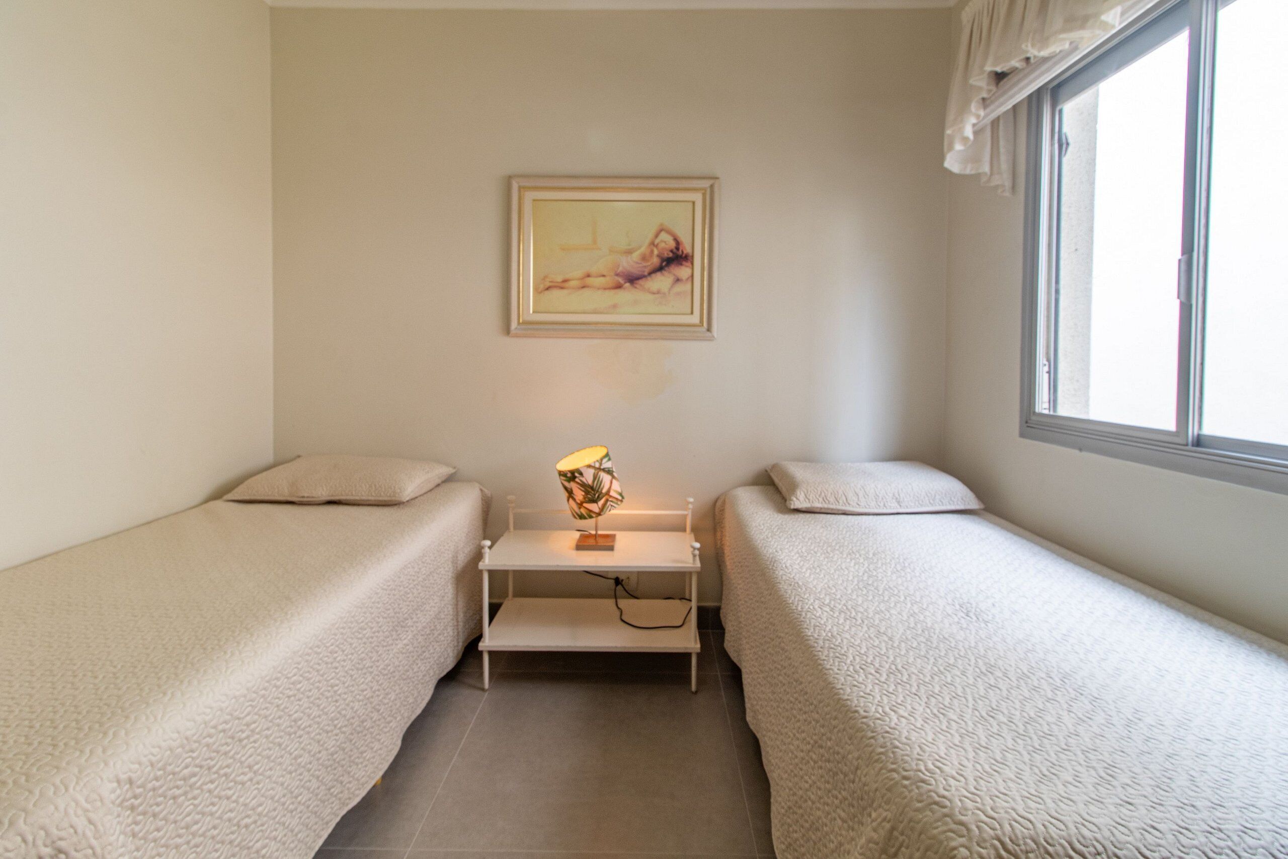 A bedroom with two single beds, a bedside table with a lamp, a framed picture on the wall, and a window with curtains.
