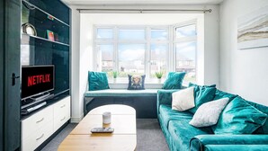 Una sala de estar con un sofá azul verdoso, una mesa de centro, un televisor sobre un mueble bajo y una ventana con vistas a casas.