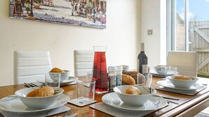 Una mesa de comedor dispuesta con platos, tazones, vasos y una botella, con una ventana y un cuadro al fondo.