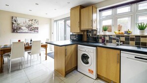 Una cocina moderna con lavadora integrada, una zona de comedor con sillas blancas y un amplio ventanal con plantas en el alféizar.
