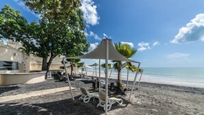 Zona de playa con sillones, una estructura con sombra y un edificio al fondo.