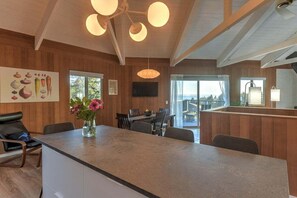 A modern kitchen with a central island, wooden walls, and a dining area with a view. - Stellar View | Panoramic Ocean Views & Hot Tub (Sea Ranch)