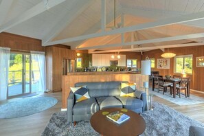 A spacious living area with a sofa, coffee table, and a dining table set for four. A kitchen with modern appliances is visible in the background. - Stellar View | Panoramic Ocean Views & Hot Tub (Sea Ranch)