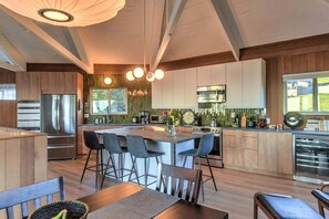 A modern kitchen with a central island, bar stools, and a dining area with a wooden table and chairs. - Stellar View | Panoramic Ocean Views & Hot Tub (Sea Ranch)