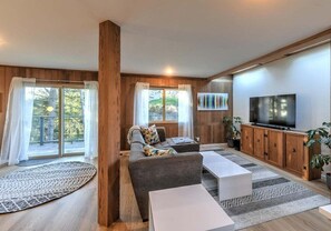 A modern living room with a sofa, coffee table, and TV, wooden walls, and large windows. - Stellar View | Panoramic Ocean Views & Hot Tub (Sea Ranch)