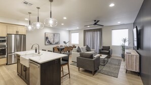 A modern kitchen with stainless steel appliances, a dining area with a table and chairs, a living room with a sofa and armchairs, and a large window with curtains. - Modern 3BR, Next to Pool, Hot Tub, Golf Views (St. George)