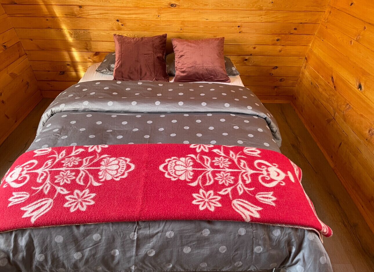 Una habitación de cabaña de madera con cama, almohadas y una manta roja estampada con flores.