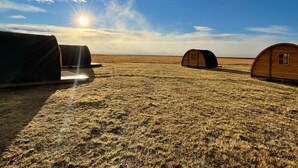 A series of wooden cabins in a vast, open field with the sun low on the horizon. - Starlight Camping Pods (Rangárþing eystra)