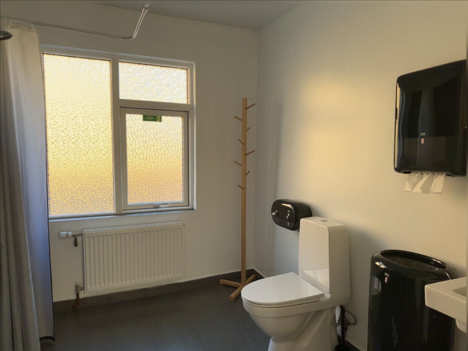 A bathroom with a toilet, a radiator, a window with frosted glass, a wooden coat rack, a black trash bin, and a wall-mounted paper dispenser.