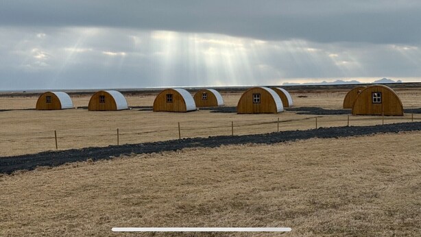 A row of wooden dome-shaped cabins in a dry, open field with a cloudy sky. - Starlight Camping Pods (Rangárþing eystra)