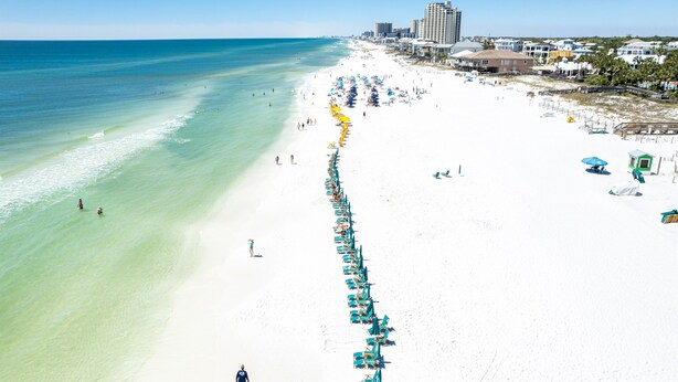 Ein Strand mit Reihen von Liegestühlen und Sonnenschirmen, einem klaren, blauen Meer und Gebäuden im Hintergrund.