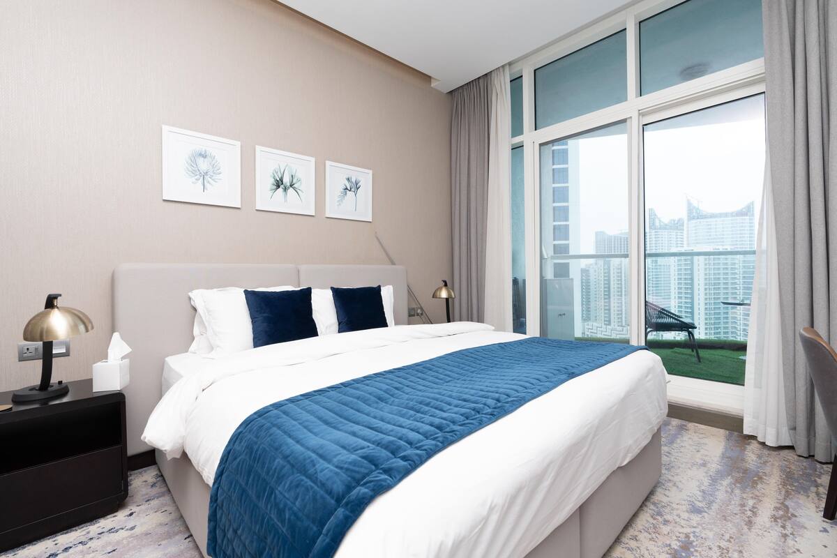 Ein modernes Hotelzimmer mit einem großen Bett, einem Nachttisch, einer Schreibtischlampe und Blick auf die Stadt durch große Fenster.