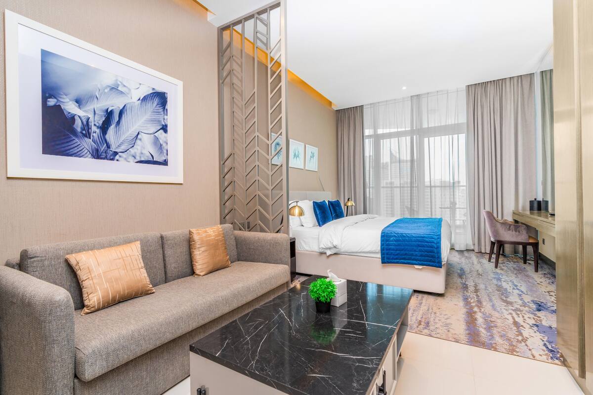 Ein modernes Hotelzimmer mit einer Couch, einem Bett mit blauer Decke, einem Schreibtisch und einem großen Fenster.