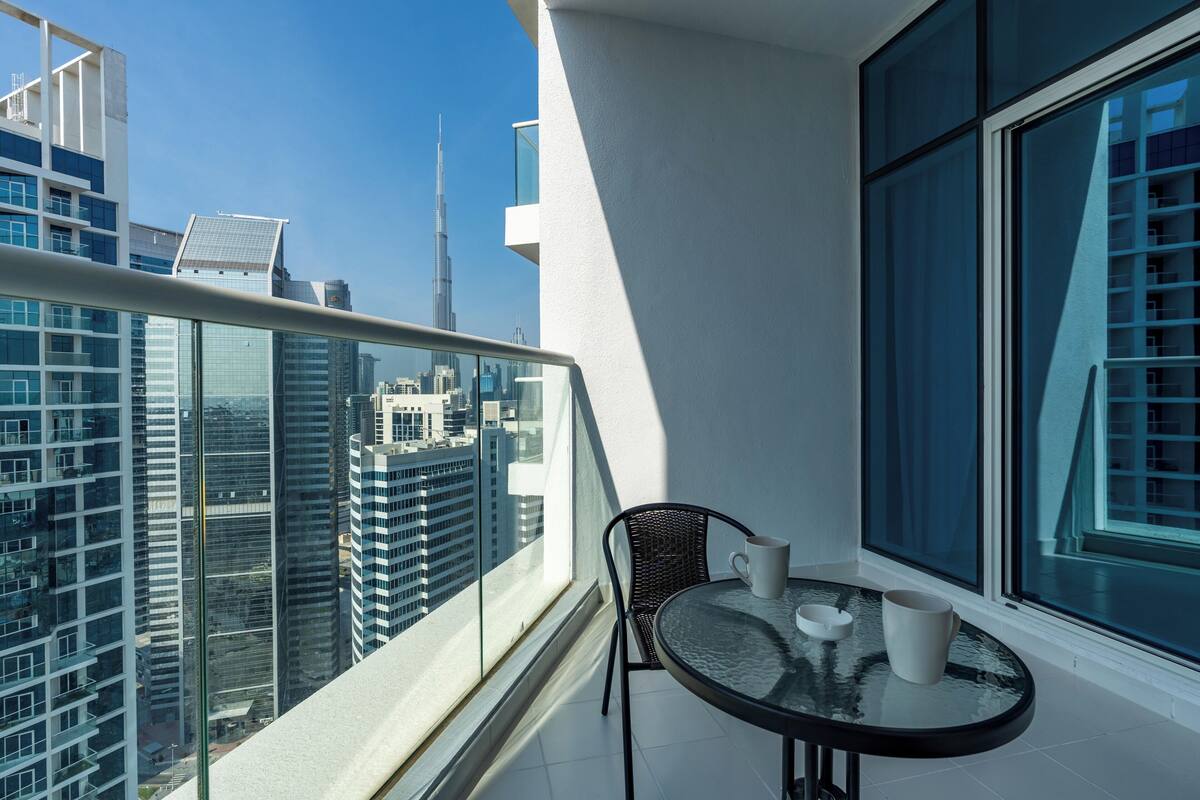 Ein Balkon mit Glastrennwand, ein Tisch mit zwei weißen Tassen und Blick auf eine Skyline, darunter ein hoher Wolkenkratzer.