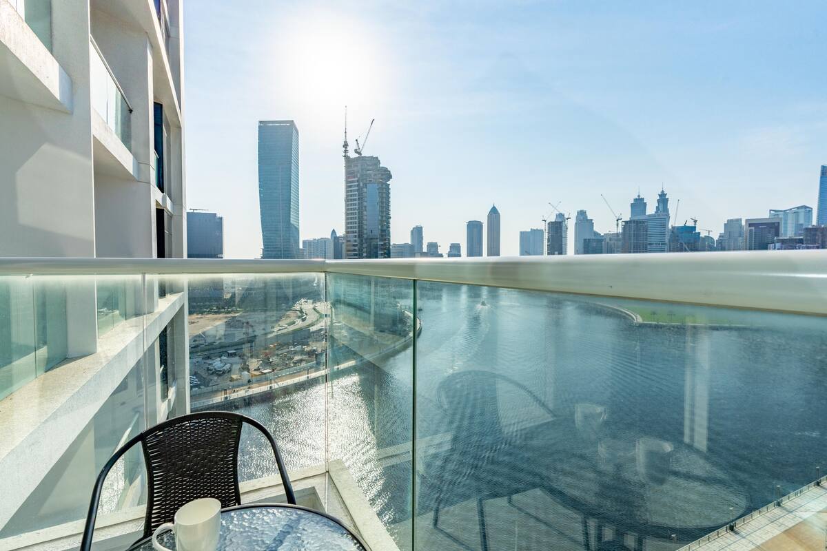 Ein Balkon mit Glasgeländer, der auf eine Stadtlandschaft und ein Gewässer blickt.