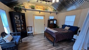 Une chambre avec un plancher en bois, un lit recouvert d’une couvre-lit à motifs, une chaise, une petite armoire et une étagère avec des plantes.