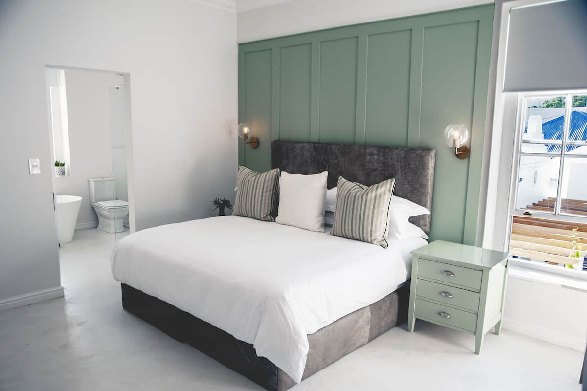 A bedroom with a large bed, a nightstand, a dresser, a window with a view, and a bathroom in the background.