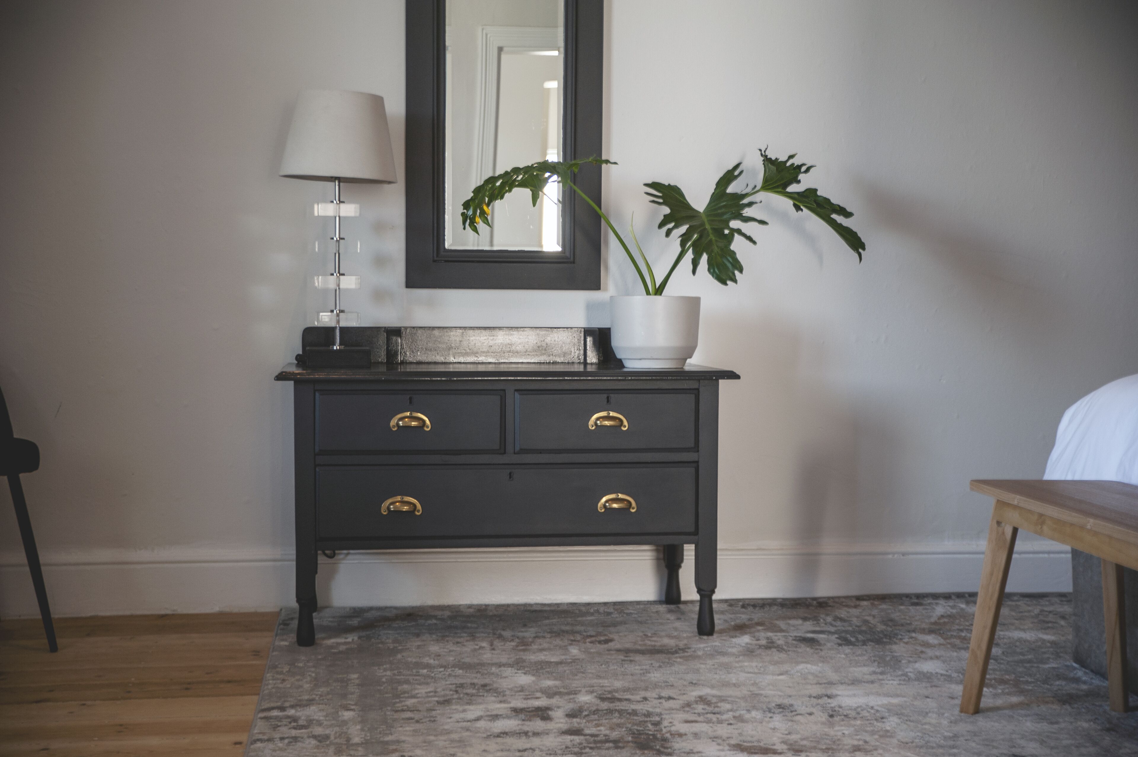 Une commode en bois foncé avec des poignées dorées, une lampe blanche et une plante en pot aux grandes feuilles.