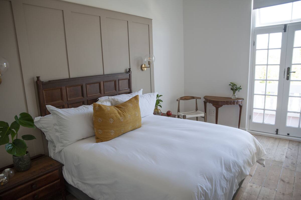 A neatly made bed with white and mustard pillows, a wooden headboard, and a small bedside table with a plant.