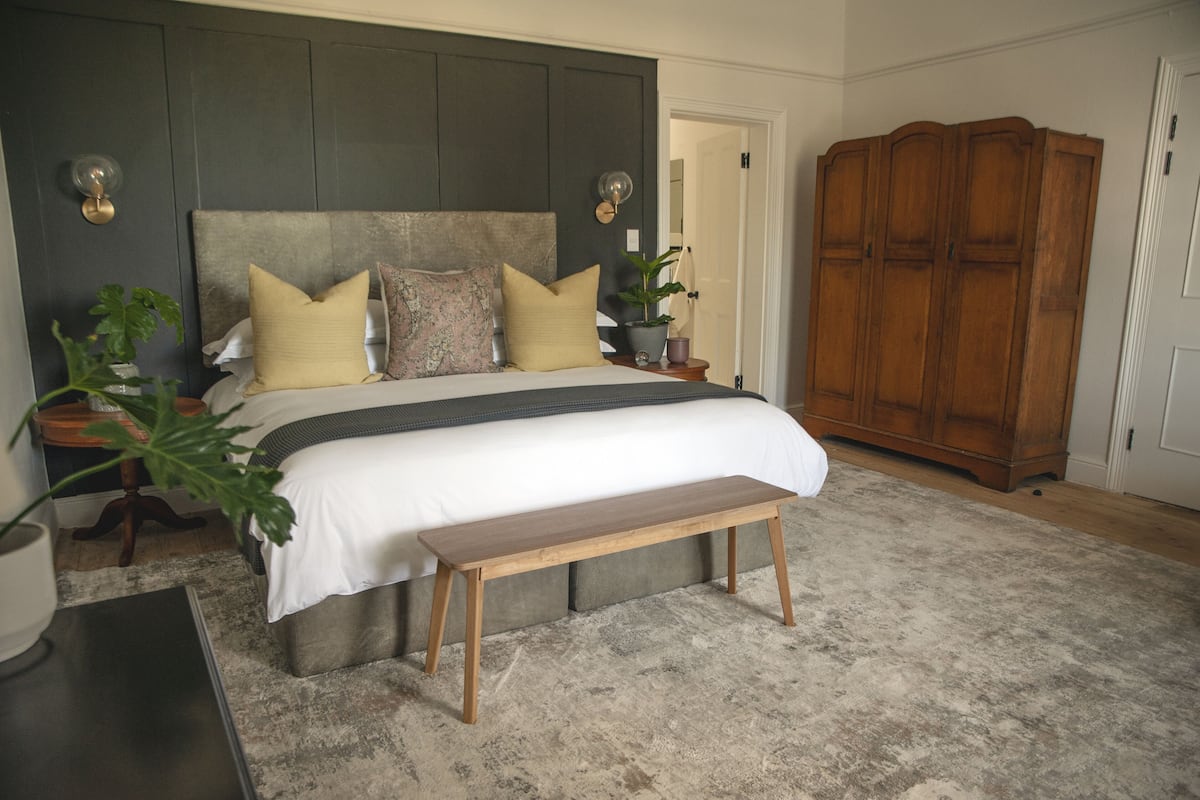 A bedroom with a large bed, a wooden bench, a wardrobe, and a nightstand with a lamp and a plant.